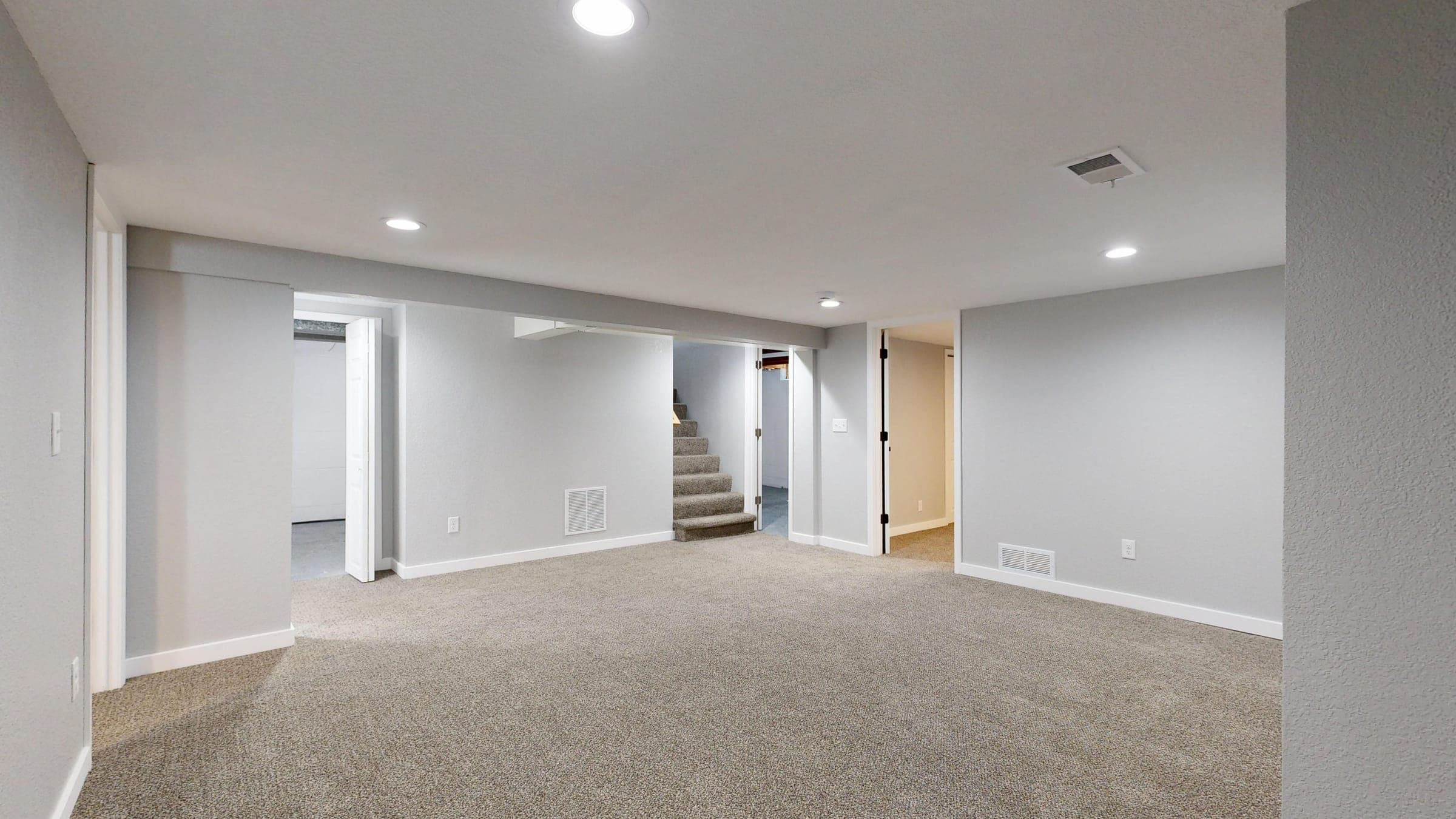 A clean, dry, well-lit finished basement with neutral painted walls, carpeted floor, and a staircase in the background.